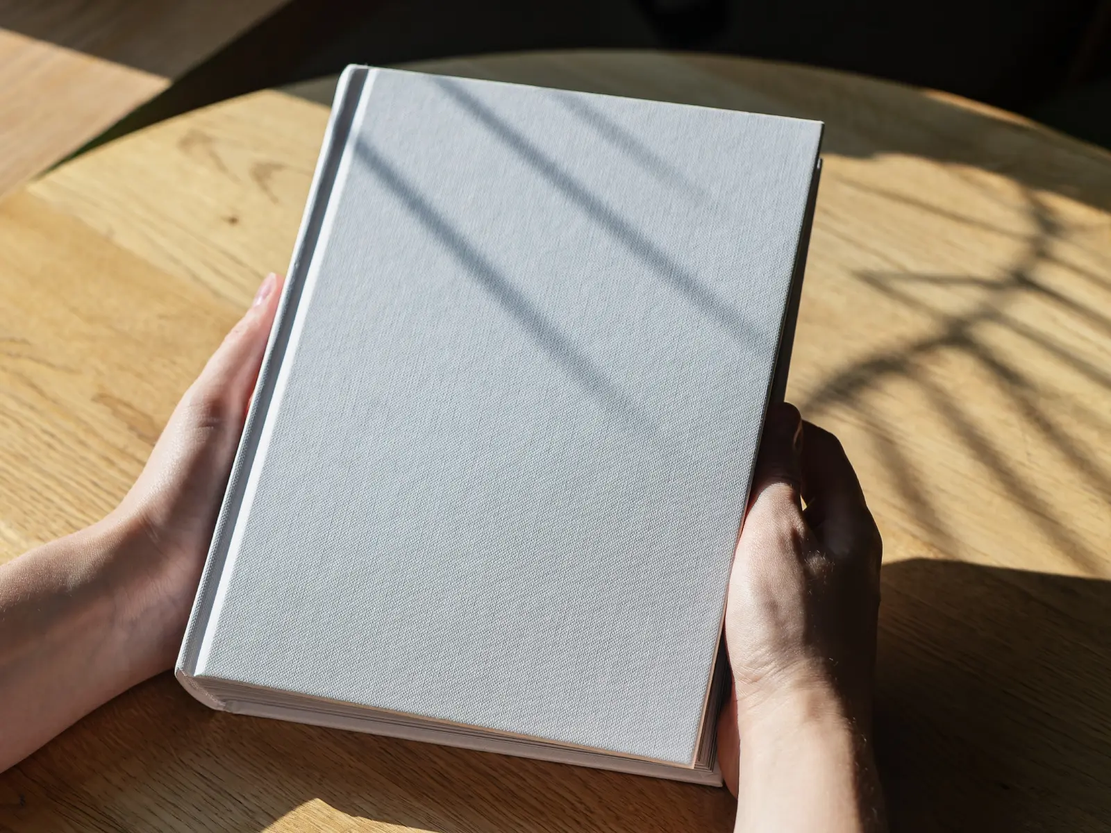 A person holds a closed, plain white hardcover book on a wooden table with sunlight casting shadows across it.