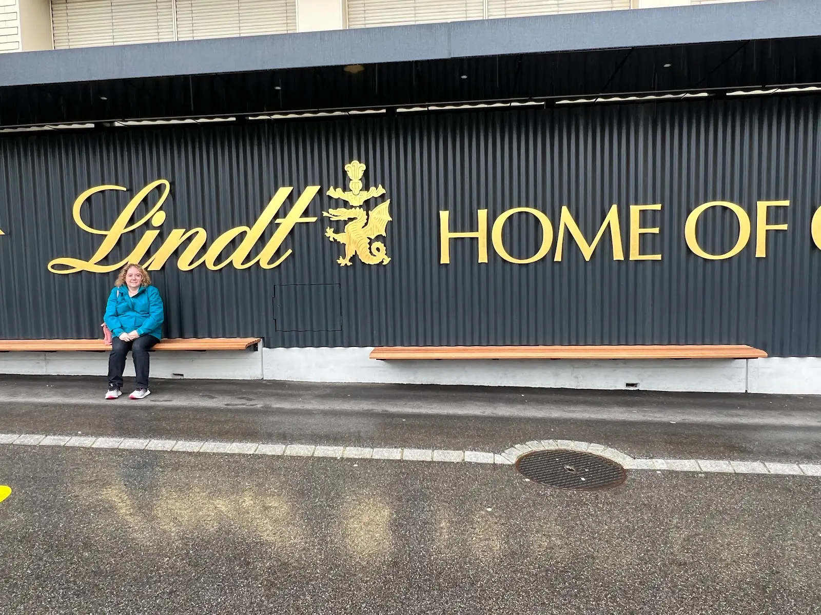 Person sitting on a bench outside the Lindt Home of Chocolate building with a large logo on the facade.
