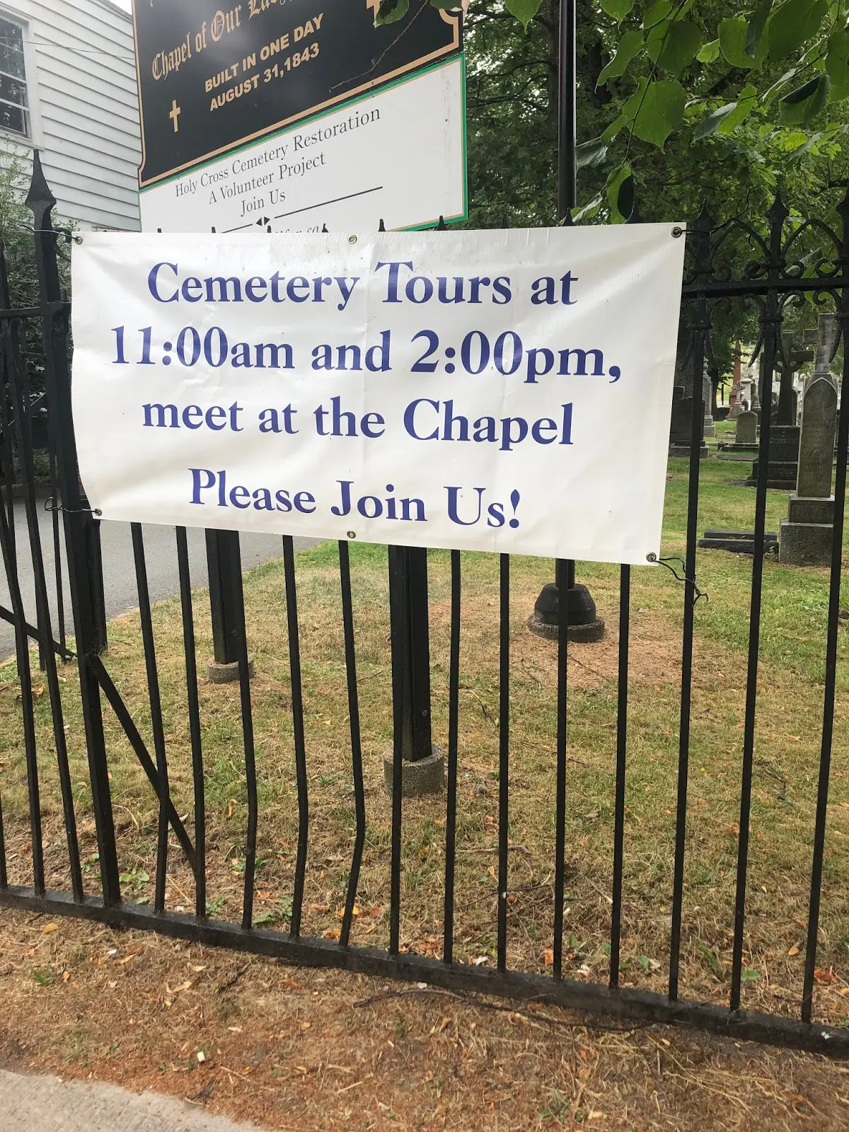 Banner on a cemetery gate reads: "Cemetery Tours at 11:00am and 2:00pm, meet at the Chapel. Please Join Us!.